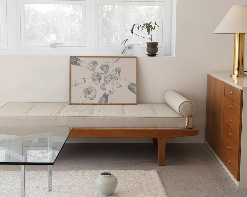 May include: A black and white line drawing of hands holding glasses of wine, framed and hanging on a wall above a light brown couch with a white cushion.