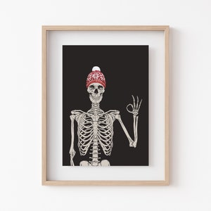Op de afbeelding: Een ingelijste kunstdruk met een illustratie van een menselijk skelet tegen een effen zwarte achtergrond. Het skelet draagt een rode gebreide muts met een wit sneeuwvlokpatroon en een witte pompon, terwijl het een ok&eacute;-handgebaar maakt.