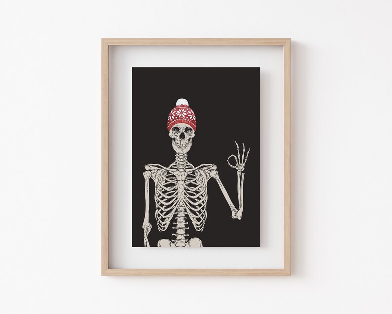 Op de afbeelding: Een ingelijste kunstdruk met een illustratie van een menselijk skelet tegen een effen zwarte achtergrond. Het skelet draagt een rode gebreide muts met een wit sneeuwvlokpatroon en een witte pompon, terwijl het een ok&eacute;-handgebaar maakt.