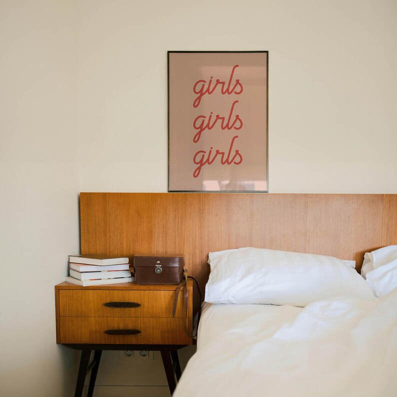 May include: A bedroom scene featuring a wooden headboard and a nightstand. A framed print with the word "girls" repeated three times hangs above the bed. The nightstand holds books and a small brown case.