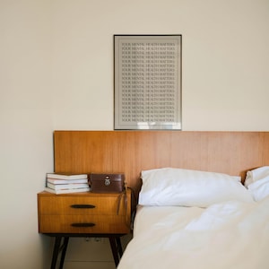 May include: A bedroom with a wooden headboard and a bedside table. A framed print with the text "YOUR MENTAL HEALTH MATTERS" is on the wall. The bedside table has books and a small brown leather case. A white bedspread and pillow are on the bed.