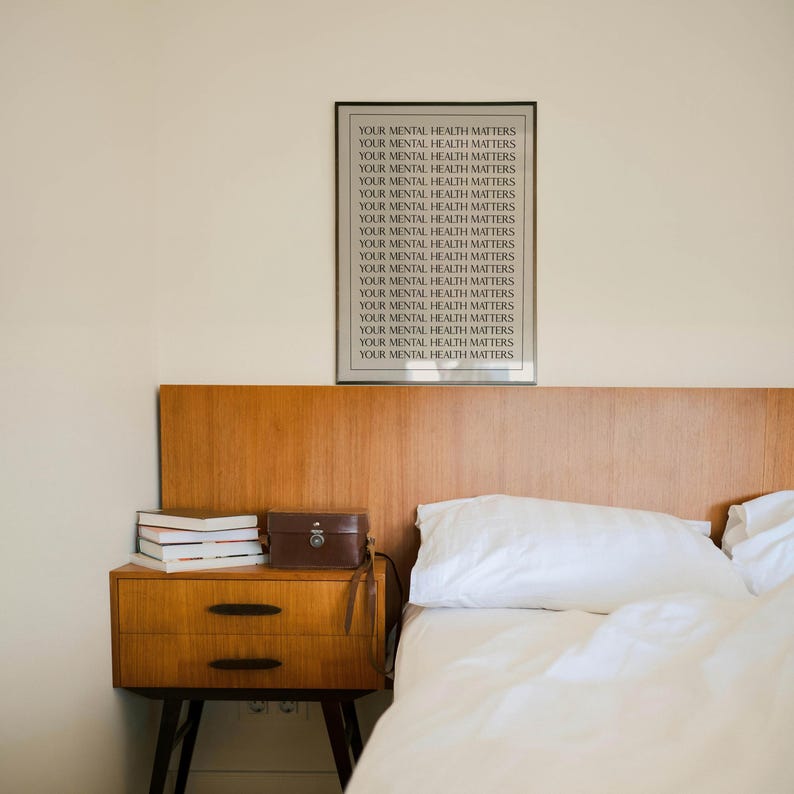 May include: A bedroom with a wooden headboard and a bedside table. A framed print with the text "YOUR MENTAL HEALTH MATTERS" is on the wall. The bedside table has books and a small brown leather case. A white bedspread and pillow are on the bed.