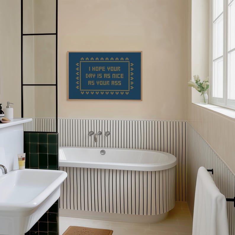 May include: A bathroom scene featuring a framed cross-stitch artwork with the text "I HOPE YOUR DAY IS AS NICE AS YOUR ASS." The artwork is framed in a wooden frame and hangs above a white bathtub. The bathroom has a neutral color palette with black and white striped accents.
