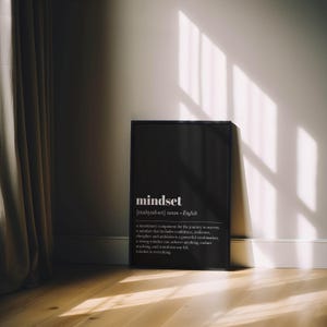 K&ouml;nnte beinhalten: Ein schwarzer gerahmter Druck mit dem Wort "mindset" in Wei&szlig; und seiner Definition. Der Druck steht an einer wei&szlig;en Wand auf einem Holzboden, mit Schatten durch Sonnenlicht.
