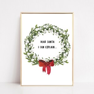 May include: A framed print featuring a watercolor wreath with green leaves, white berries, and a red bow. The text "DEAR SANTA I CAN EXPLAIN..." is centered within the wreath. The frame is gold.