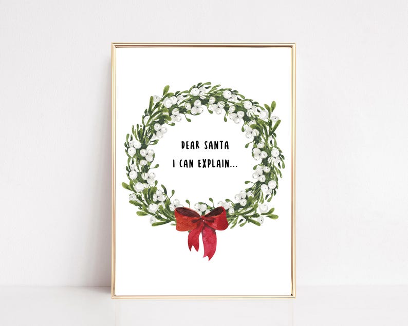 May include: A framed print featuring a watercolor wreath with green leaves, white berries, and a red bow. The text "DEAR SANTA I CAN EXPLAIN..." is centered within the wreath. The frame is gold.