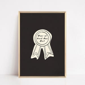 Op de afbeelding: Een ingelijste print met een zwarte achtergrond en een gouden lijst. De print toont een crèmekleurig lint met de woorden "Doer of Hard Ass Shit" in een cirkelvormig ontwerp.