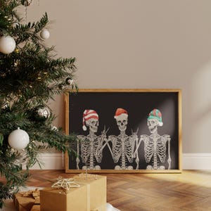 May include: A framed horizontal print on a black background featuring three white skeletons wearing festive hats. The left skeleton wears a red and white striped hat, the centre wears a red Santa hat, and the right wears a green and red striped hat.