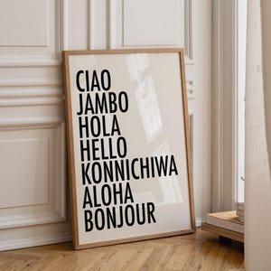 May include: A framed print with a cream-colored background and black text. The text includes the words "CIAO, JAMBO, HOLA, HELLO, KONNICHIWA, ALOHA, BONJOUR". The frame is made of light-colored wood, and the print is leaning against a white wall.