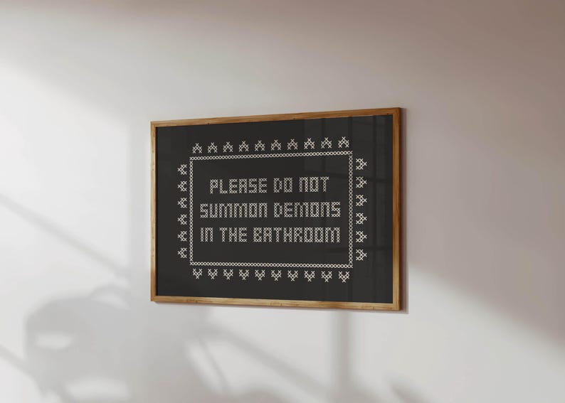 May include: A framed black cross-stitch artwork with the white text "PLEASE DO NOT SUMMON DEMONS IN THE BATHROOM". The piece is in a wooden frame and hangs on a white wall. The text is centered within a white border, creating a humorous message.