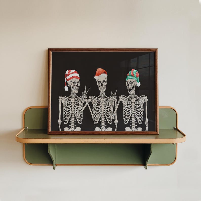 May include: A framed horizontal print featuring three white skeletons against a black background. Each skeleton wears a different coloured holiday hat: red and white striped, solid red, and green and red striped. The skeletons are making hand gestures.