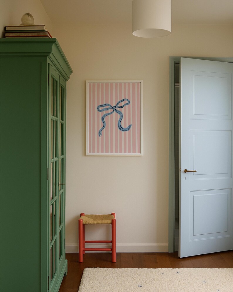 Pode incluir: Uma impress&atilde;o de arte emoldurada com um la&ccedil;o azul desenhado &agrave; m&atilde;o, centrado num fundo de riscas verticais cor-de-rosa claro e branco. A obra de arte est&aacute; exposta numa parede lisa entre um arm&aacute;rio verde e uma porta azul-clara.