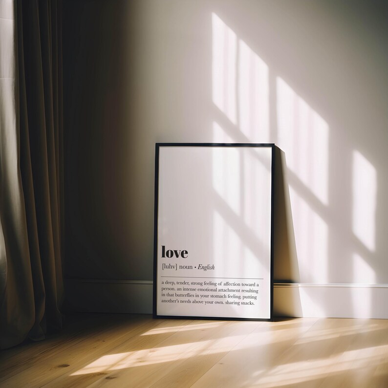 May include: A framed print with the definition of the word "love" in black text on a white background. The print is leaning against a white wall on a wooden floor, with sunlight casting shadows.