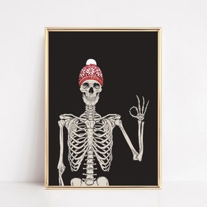 Op de afbeelding: Een ingelijste kunstdruk met een menselijk skelet tegen een effen zwarte achtergrond. Het skelet draagt een rode gebreide muts met een wit sneeuwvlokpatroon en een witte pompon, terwijl het een ok&eacute;-gebaar maakt met de linkerhand.