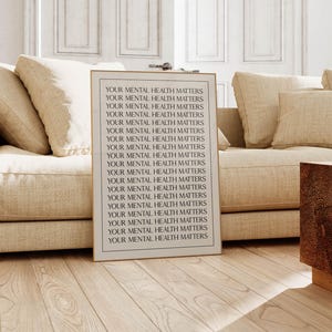 May include: A framed print with the text "YOUR MENTAL HEALTH MATTERS" repeated in black. The print is in a living room, leaning against a beige sofa. The room has light-coloured wooden floors and a wooden side table. The overall aesthetic is neutral.