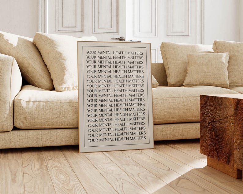 May include: A framed print with the text "YOUR MENTAL HEALTH MATTERS" repeated in black. The print is in a living room, leaning against a beige sofa. The room has light-coloured wooden floors and a wooden side table. The overall aesthetic is neutral.