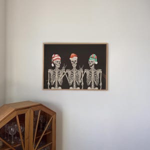 May include: A framed horizontal wall art print featuring three skeletons against a black background. Each skeleton wears a different holiday hat: one red and white striped, one solid red, and one green and red. The skeletons are making hand gestures.