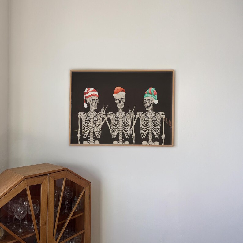 May include: A framed horizontal wall art print featuring three skeletons against a black background. Each skeleton wears a different holiday hat: one red and white striped, one solid red, and one green and red. The skeletons are making hand gestures.
