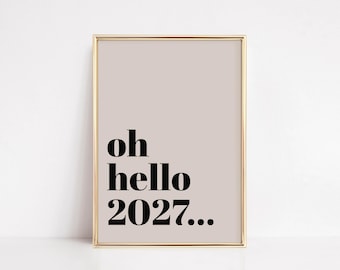 May include: A framed print with a gold-colored border. The print has a light tan background with the words "oh hello 2027..." in black, bold font. The artwork is a minimalist design.