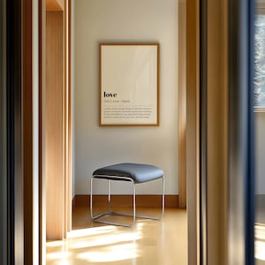 May include: A modern interior scene featuring a framed print with the word "love" and its definition. Below, a gray, square-topped stool with a chrome frame sits on a light wood floor. The room is bathed in natural light.