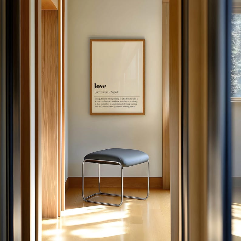May include: A modern interior scene featuring a framed print with the word "love" and its definition. Below, a gray, square-topped stool with a chrome frame sits on a light wood floor. The room is bathed in natural light.