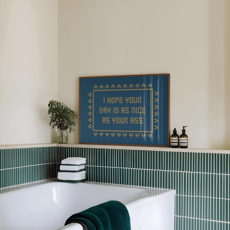 May include: A framed cross-stitch artwork with the text "I HOPE YOUR DAY IS AS NICE AS YOUR ASS" in gold thread on a blue background. The artwork is displayed in a bathroom setting, above a bathtub with green tile.
