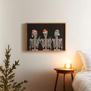 May include: A framed rectangular wall art print featuring three skeletons against a black background. Each skeleton wears a different holiday-themed hat: one with red and white stripes, one solid red, and one green and red. They are making hand gestures.