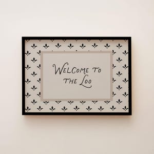 May include: A framed sign with the text "Welcome to the Loo" in elegant script. The sign has a black frame and a patterned background with black floral motifs on a white background. The inner border is a light gray.