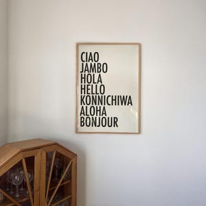 May include: A framed print on a white background displays the words "Ciao, Jambo, Hola, Hello, Konnichiwa, Aloha, Bonjour" in a black, bold font. The frame is a light brown color. A wooden cabinet with glass doors is in the lower left corner.