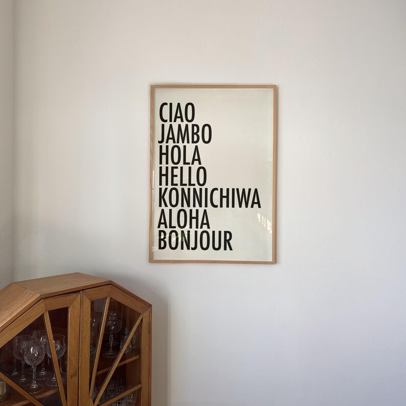 May include: A framed print on a white background displays the words "Ciao, Jambo, Hola, Hello, Konnichiwa, Aloha, Bonjour" in a black, bold font. The frame is a light brown color. A wooden cabinet with glass doors is in the lower left corner.