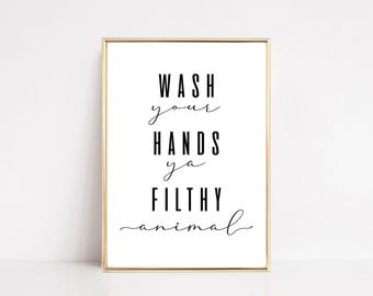 May include: A rectangular wall art print in a thin gold frame featuring black text on a white background that reads: WASH your HANDS ya FILTHY animal. The typography alternates between a bold sans-serif font and a delicate cursive script.