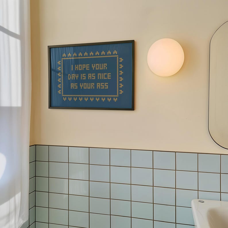 May include: Framed wall art with the text "I HOPE YOUR DAY IS AS NICE AS YOUR ASS" in a cross-stitch style. The artwork is in a black frame and hangs on a cream-colored wall above blue tiles. A round light fixture is to the right.