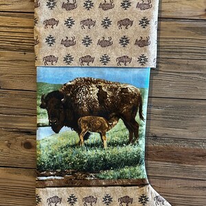 May include: A brown and tan Christmas stocking with a buffalo and calf design. The stocking has a brown and tan buffalo print with a blue background and a brown and tan buffalo print on the cuff.