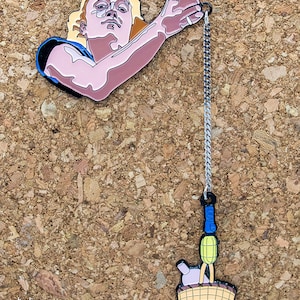 May include: An enamel pin featuring a cartoon man holding a basket with a bottle and a yellow object hanging from it. The pin is attached to a silver chain.