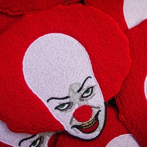 May include: A close-up of a red embroidered patch featuring a creepy clown face with a white background. The clown has a red nose, white teeth, and a sinister smile.