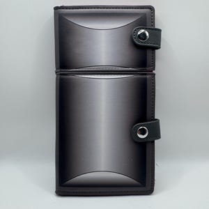 May include: A two-section, dark gray wallet with a gradient design. Each section has a curved top and a small, dark gray strap with a silver snap closure. The wallet is rectangular and appears to be made of leather.