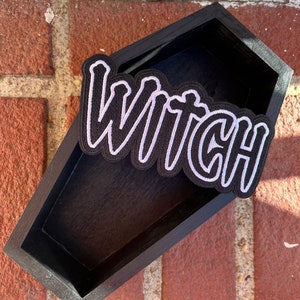 Peut inclure: Une bo&icirc;te en bois noire en forme de cercueil avec un patch brod&eacute; blanc qui dit "Witch" dessus.