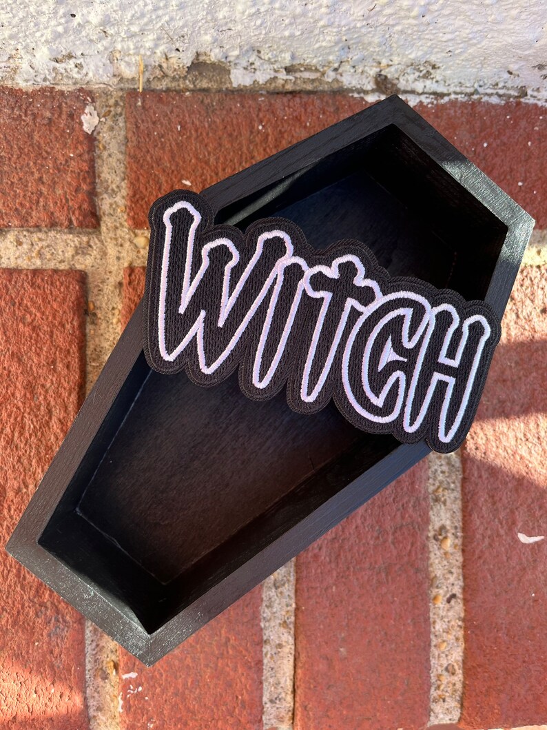 Peut inclure: Une bo&icirc;te en bois noire en forme de cercueil avec un patch brod&eacute; blanc qui dit "Witch" dessus.