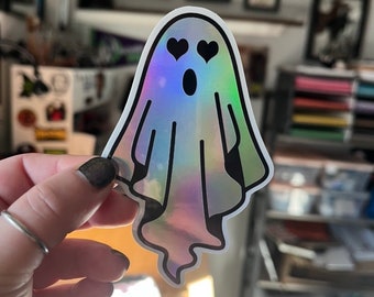 May include: A holographic ghost sticker with black outline and googly eyes. The ghost has two black hearts for pupils.