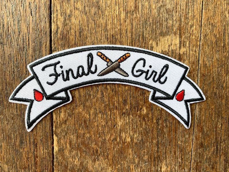 May include: White embroidered patch with a black banner that reads "Final Girl" with two crossed knives in the center. The banner has red blood drops on each end.