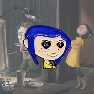 May include: Enamel pin featuring a stylized image of a character with blue hair and heart-shaped eyes. The character is wearing a yellow shirt and has freckles. The pin is set against a blurred background with other characters.