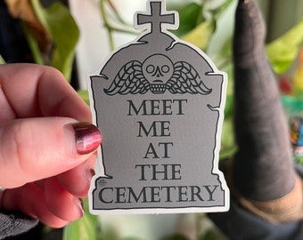 May include: A gray tombstone sticker with a skull and wings design. The sticker says "Meet Me At The Cemetery".
