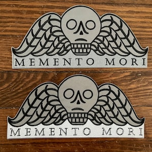 Può includere: Due toppe ricamate argento e nero con un designo di teschio e ali. Il testo "Memento Mori" è stampato sotto il teschio e le ali.