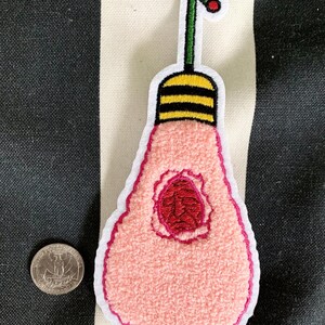 Peut inclure: Un patch en forme d'ampoule rose avec un centre rouge et une tige verte avec un point rouge sur le dessus. L'ampoule a une bande jaune et noire ray&eacute;e autour du haut.