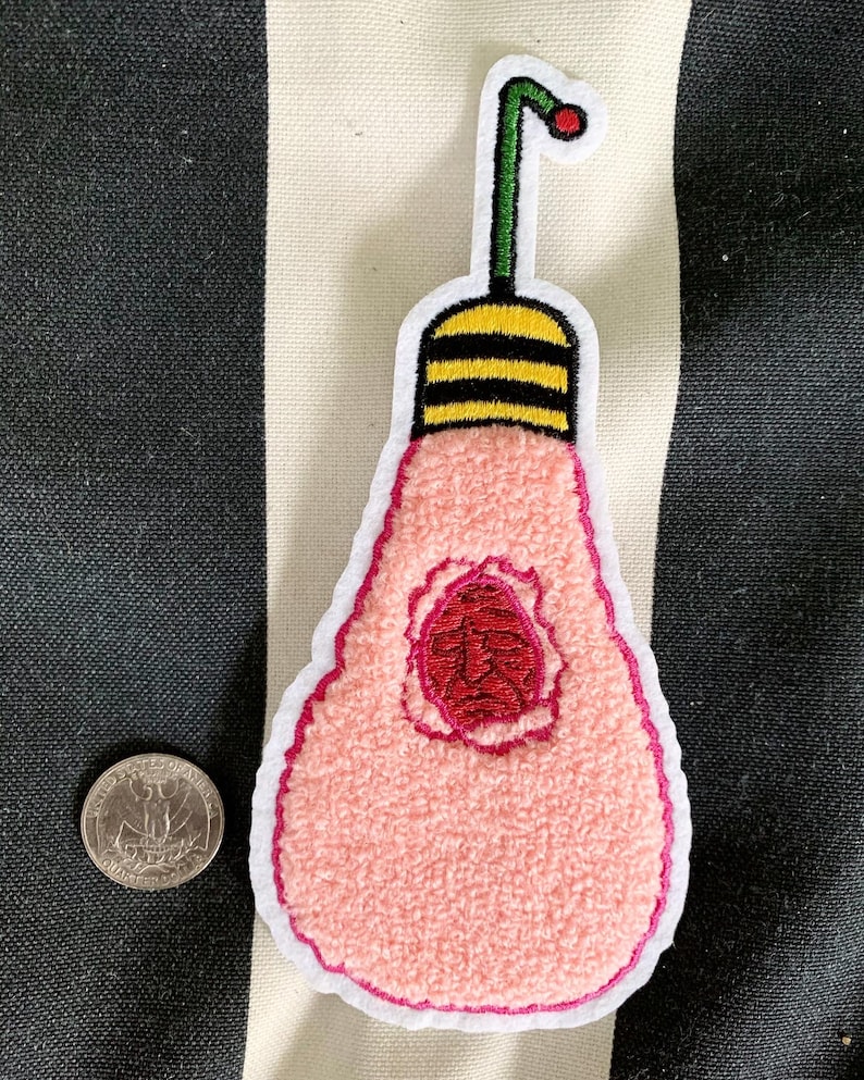 Peut inclure: Un patch en forme d'ampoule rose avec un centre rouge et une tige verte avec un point rouge sur le dessus. L'ampoule a une bande jaune et noire ray&eacute;e autour du haut.