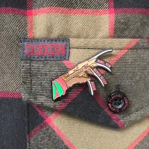 Peut inclure: Une épingle en émail noir et rouge représentant une main tenant un couteau, avec le texte "Nine, Ten, Never Sleeps Again". L'épingle est fixée à une chemise à carreaux.