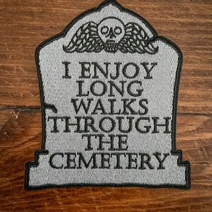 May include: A gray embroidered patch shaped like a tombstone with a skull and wings at the top. The text on the patch reads "I enjoy long walks through the cemetery."