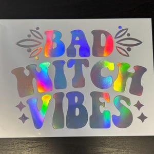 May include: A holographic sticker with the text "BAD WITCH VIBES" in a rainbow iridescent color.