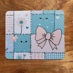 May include: Square coaster with a patchwork design featuring a large pink bow, flowers, stars, and hearts. The coaster has a light blue, pink, and white color scheme, with a stitched border.