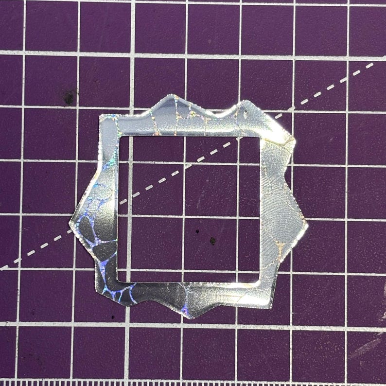May include: A silver-colored, irregularly shaped frame with a square opening. The frame has a textured, iridescent surface with a pattern of blue and black. The background is a purple grid.